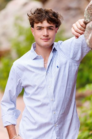 Young man in blue striped shirt leaning against rock outdoors, smiling confidently in natural setting.