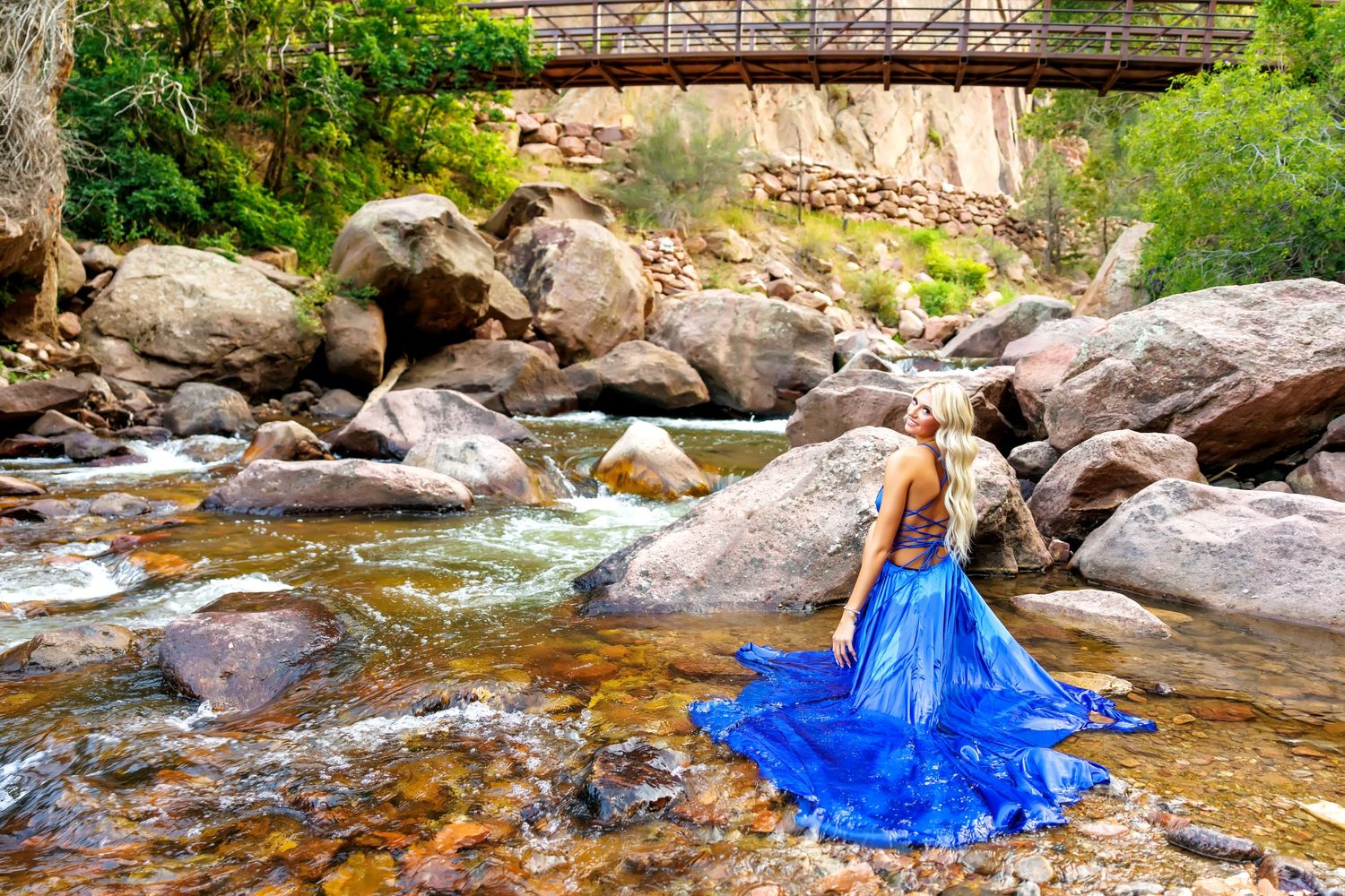 Woman in blue gown poses on rocks by a flowing stream beneath a rustic wooden bridge in a canyon.