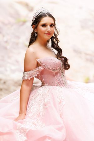 Young woman in pink off-shoulder ball gown with lace details and crystal tiara poses elegantly outdoors.