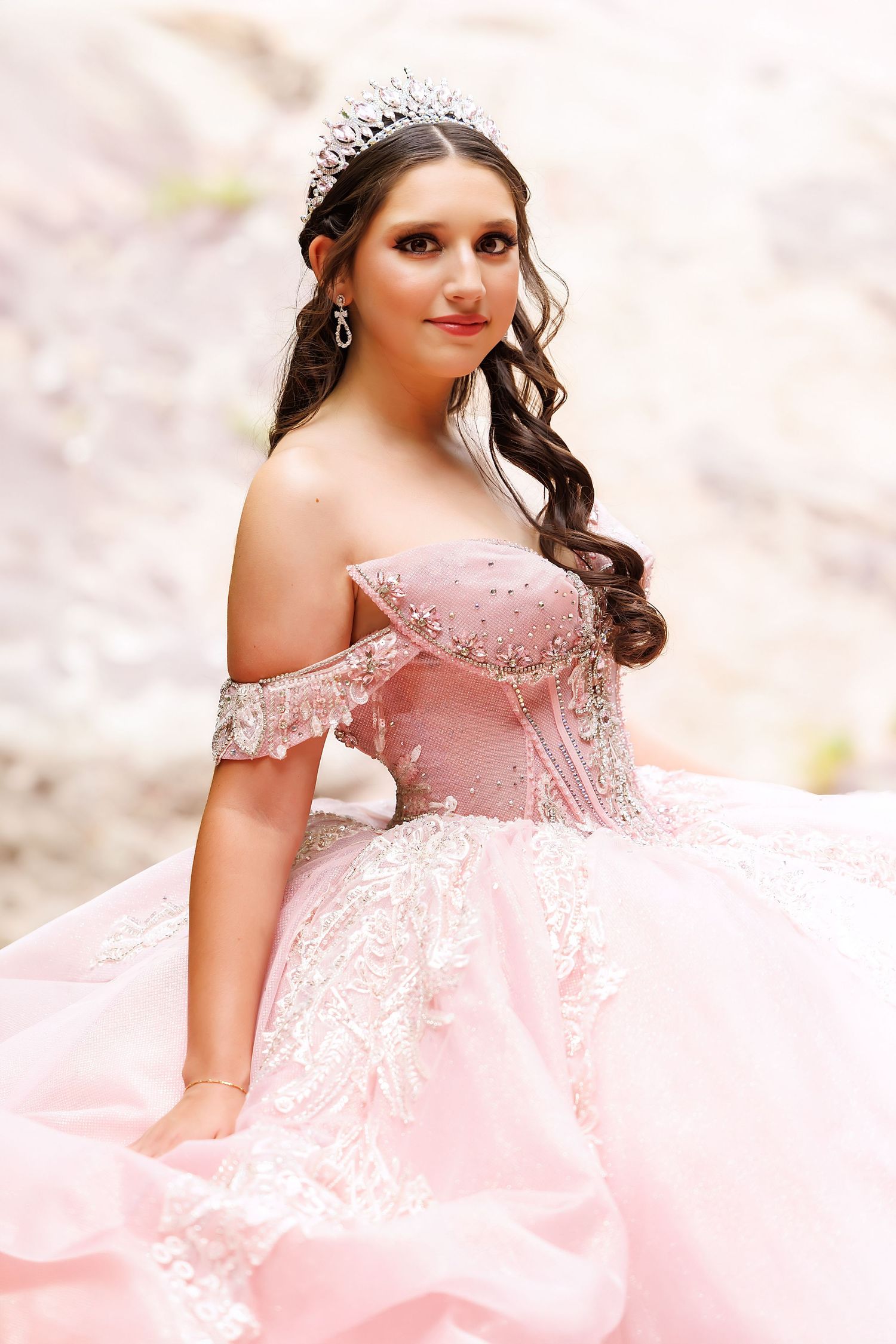 Young woman in pink off-shoulder ball gown with lace details and crystal tiara poses elegantly outdoors.