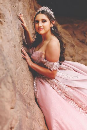 Young woman in pink off-shoulder ball gown and tiara poses against rocky wall for quinceañera portrait.
