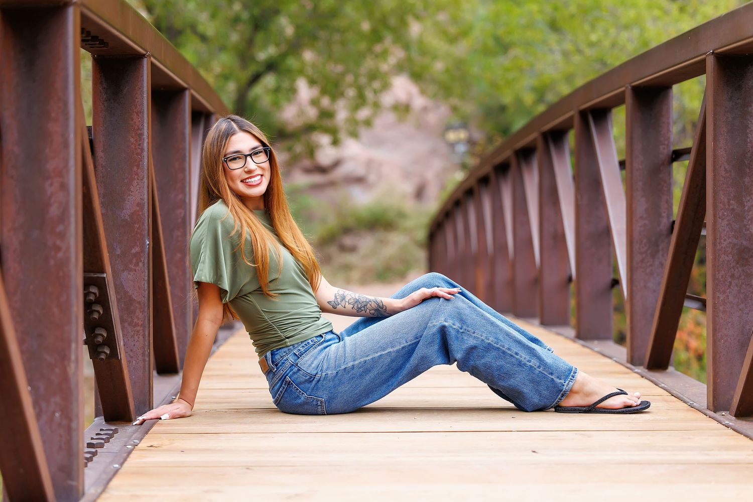 Young woman with glasses and tattoos sitting on a wooden bridge, smiling, wearing jeans and a green t-shirt.
