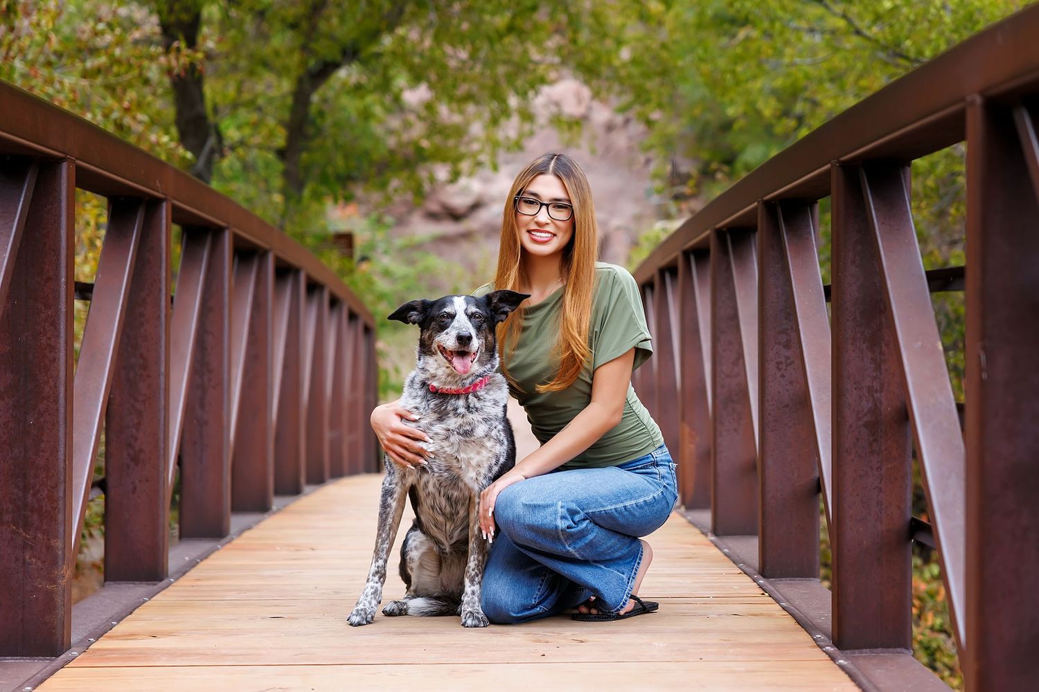 Young woman with glasses kneeling beside a spotted dog on a wooden bridge surrounded by lush green trees.