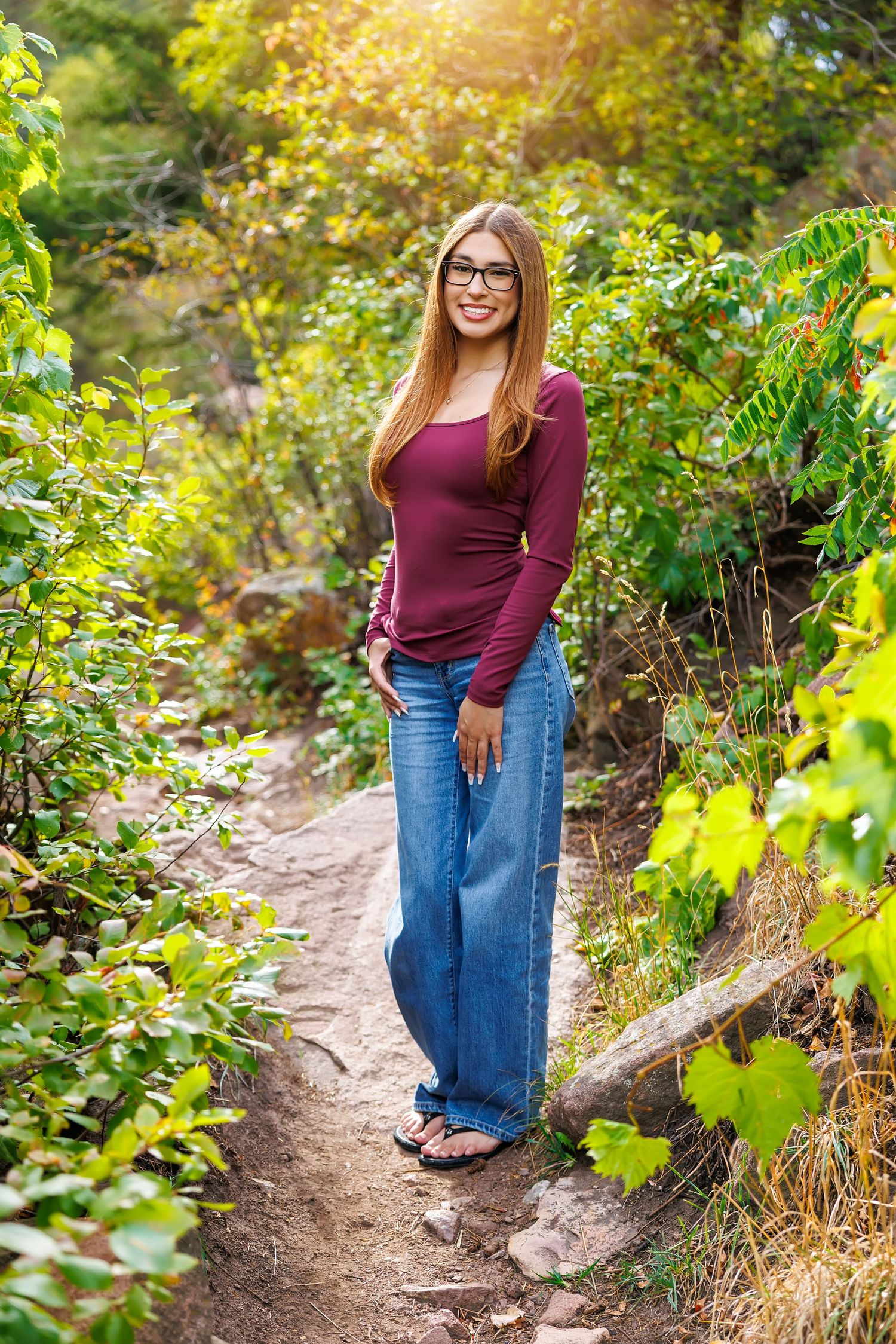 Young woman in glasses, burgundy top, and wide-leg jeans smiling on a nature trail surrounded by lush green foliage.