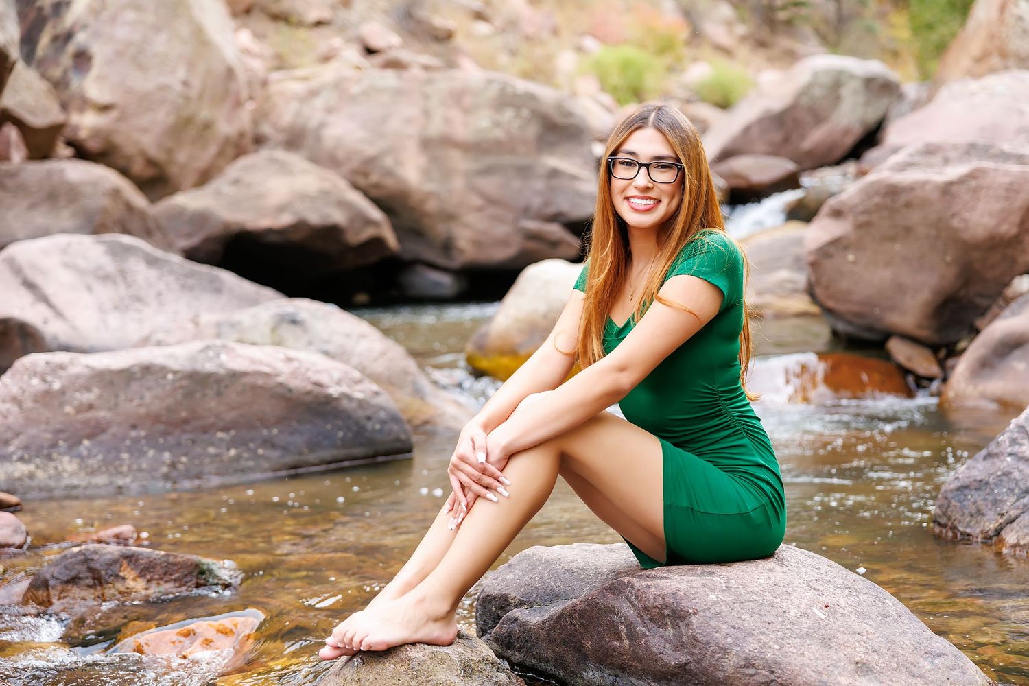 Young woman in green dress and glasses sitting barefoot on rocks by a shallow stream in nature.