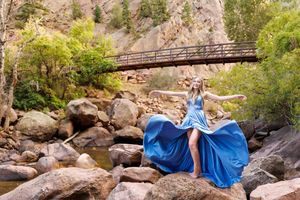 Woman in flowing blue gown poses on rocky riverbank with wooden bridge and mountain backdrop.
