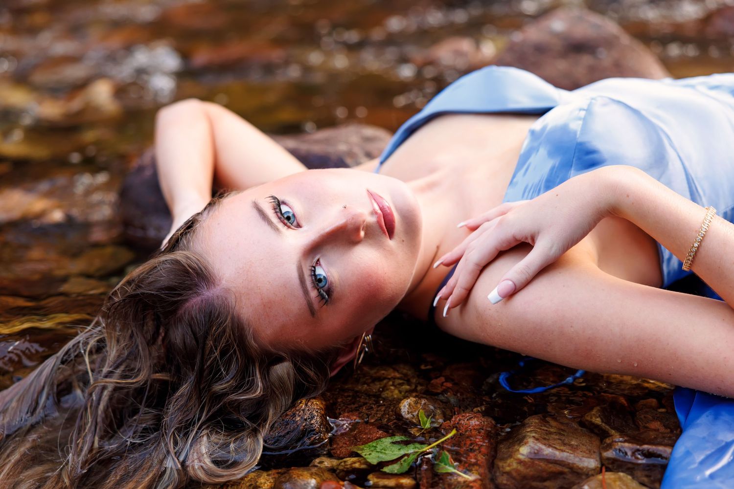 Young woman in blue dress lying on rocky shallow stream, hair spread out, gazing upward in serene outdoor portrait.