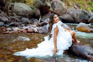 Bridal portrait of woman in white gown sitting gracefully among river rocks.