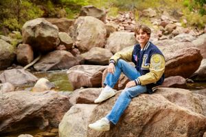 A casual portrait sitting on rocky mountain terrain wearing varsity jacket and jeans.