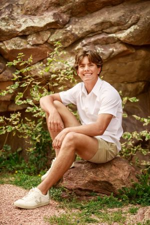 A relaxed outdoor portrait sitting on rock formation in white shirt and khaki shorts.