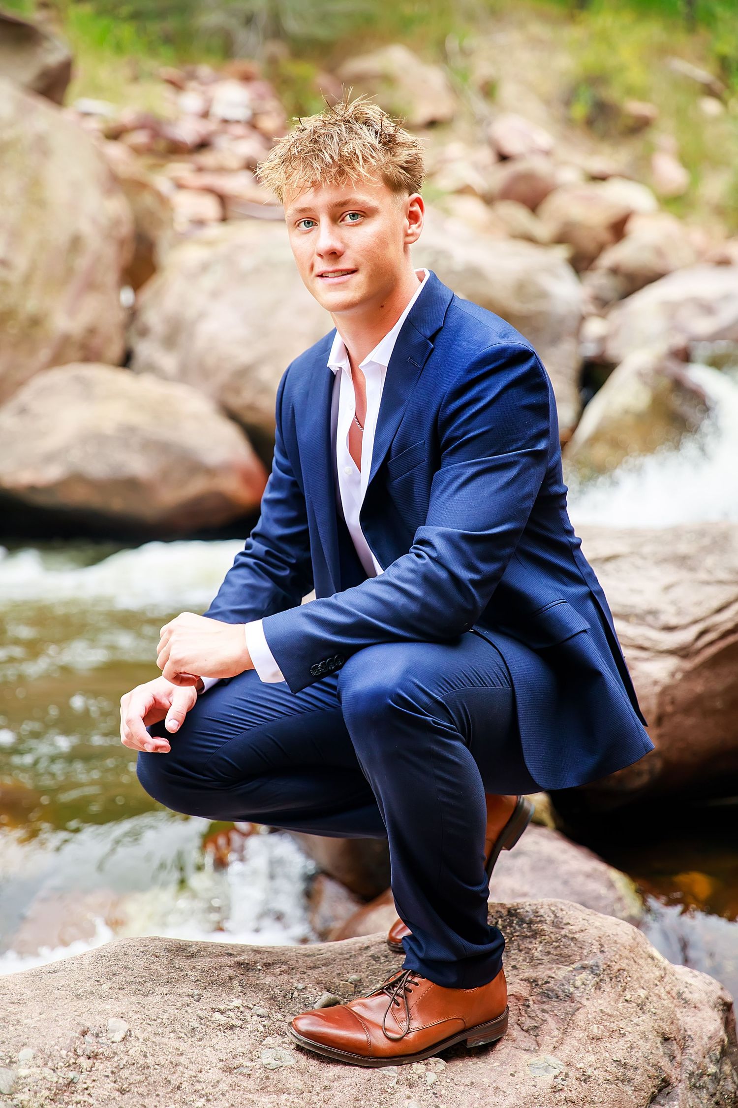 Person in navy blue suit poses on rocks near flowing stream in outdoor portrait session.