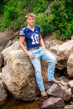 A person in a blue football jersey and light jeans sits casually on large rocks surrounded by lush green foliage.