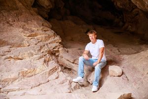 A person explores a rocky cave formation while wearing casual clothing and sitting among large boulders and crevices.
