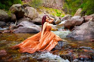 A flowing orange dress creates an ethereal effect against rocky stream beds and cascading water in a forest setting.