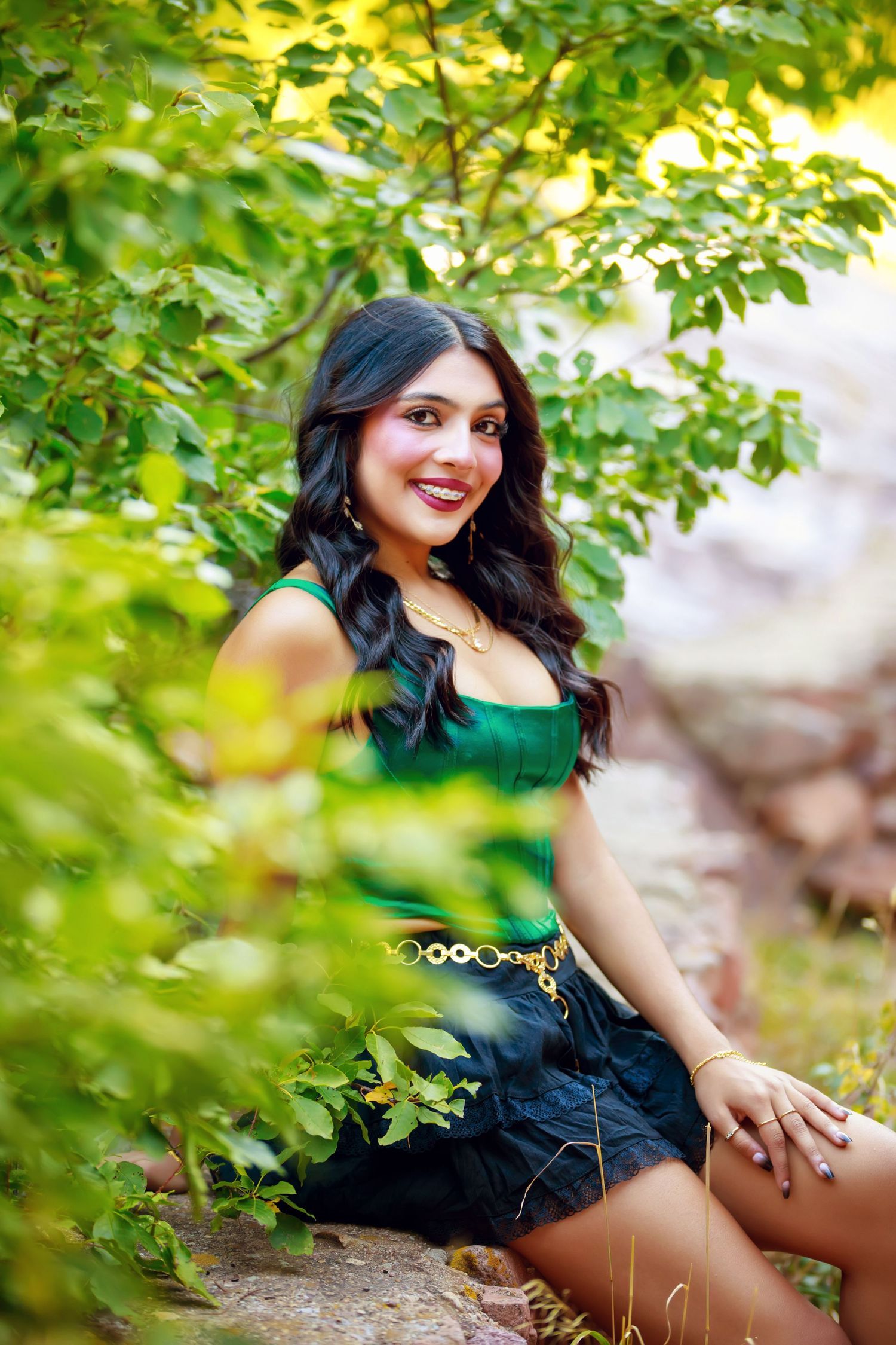 A figure in a black dress poses outdoors among green foliage in a natural setting during a photo session.