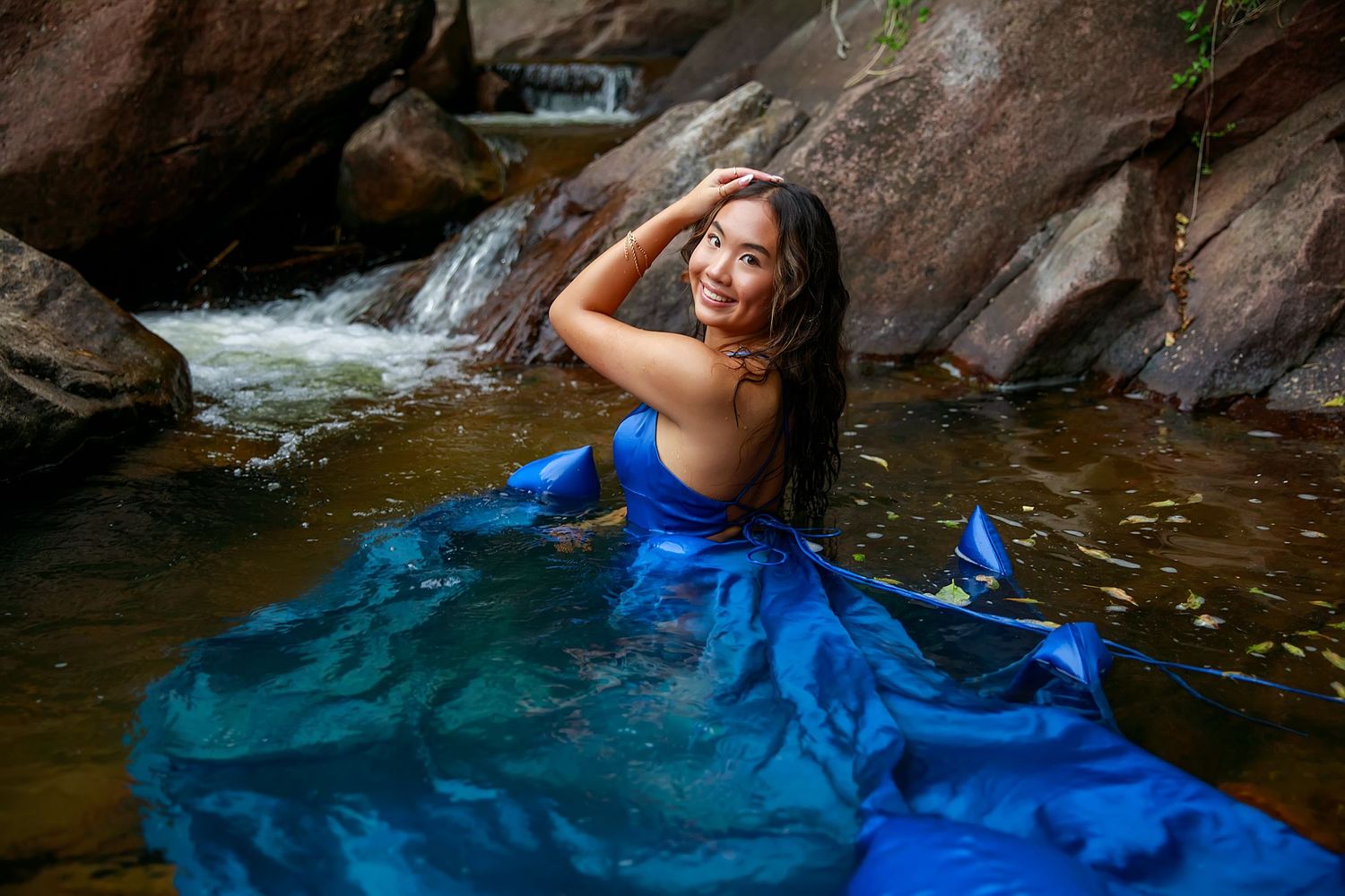 A person in flowing blue dress poses in a mountain stream with waterfall.