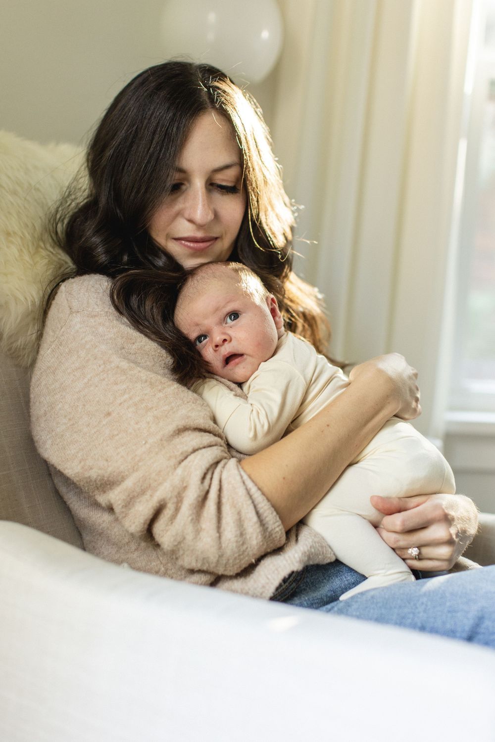 Mother tenderly cradling wide-eyed newborn baby while seated in cozy chair at home.