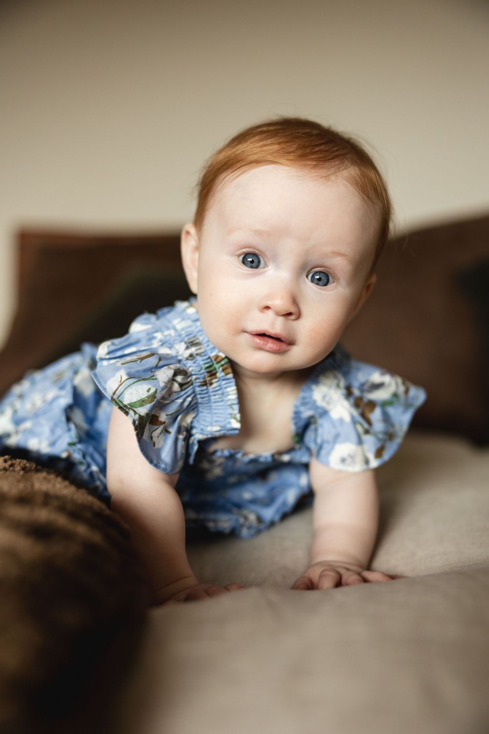Cute red-haired baby girl in blue floral dress doing tummy time on a beige couch.