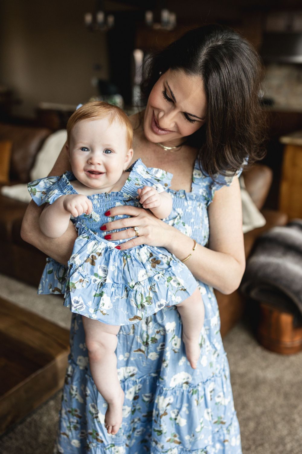 Smiling mother in blue floral dress holding happy red-haired baby girl in matching outfit indoors.