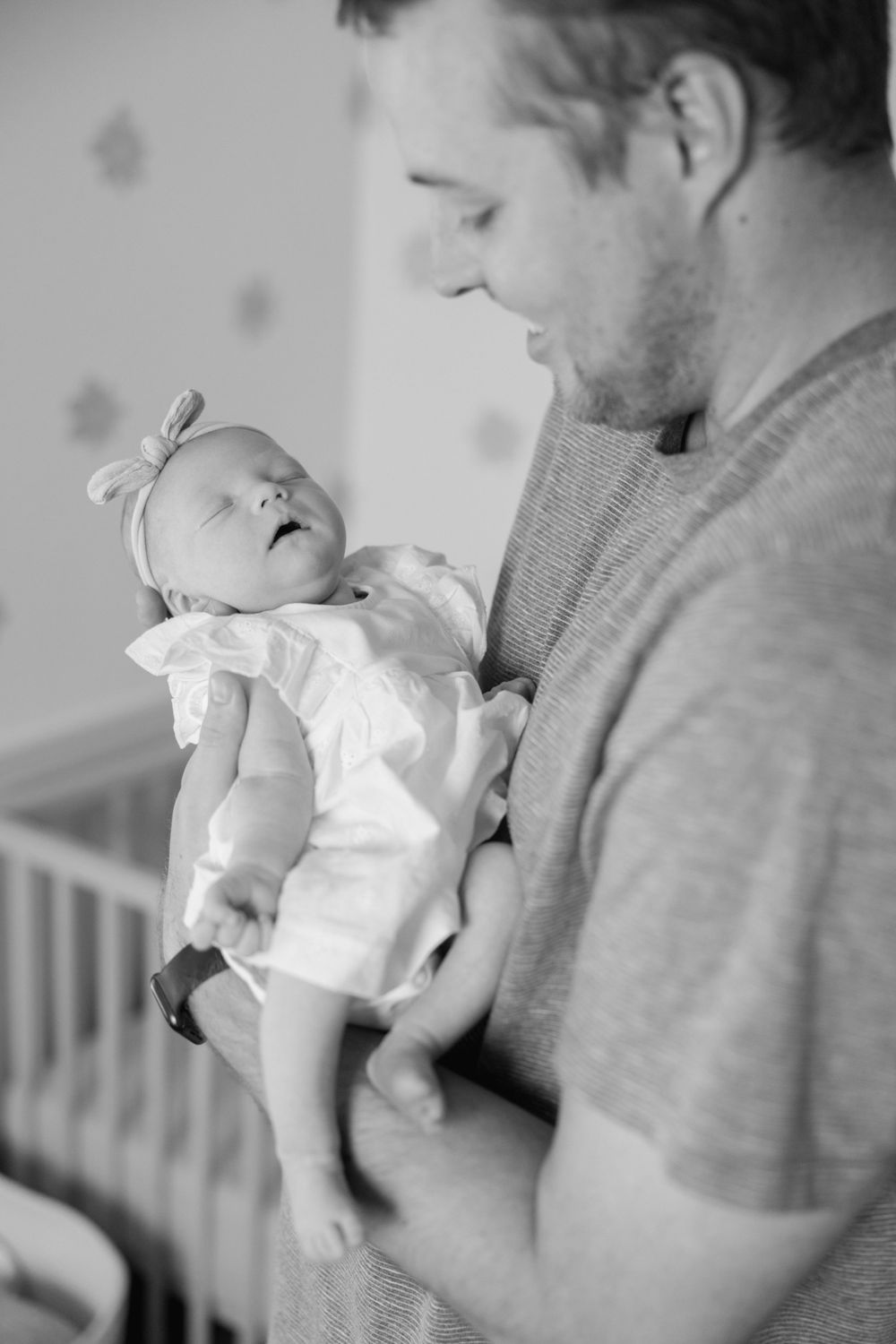 Black and white photo of a father cradling his sleeping newborn baby girl in a nursery.