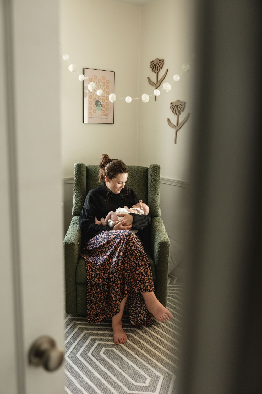 New mother sitting in green glider chair holding her newborn baby in a cozy nursery.
