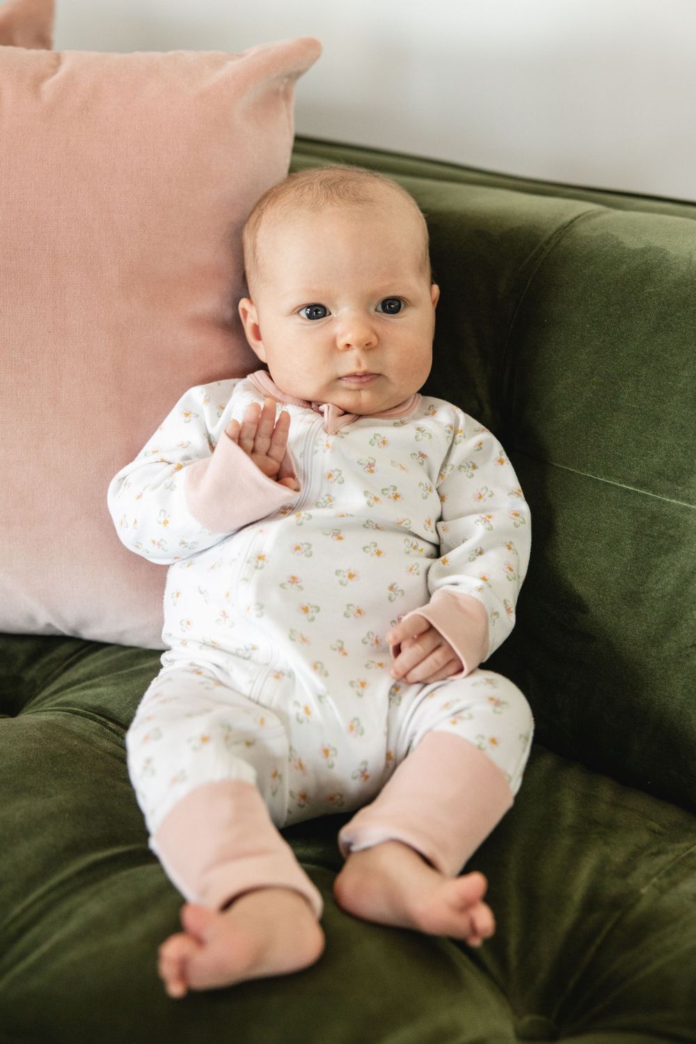 Adorable baby in floral onesie sits propped against pink pillow on green velvet sofa.