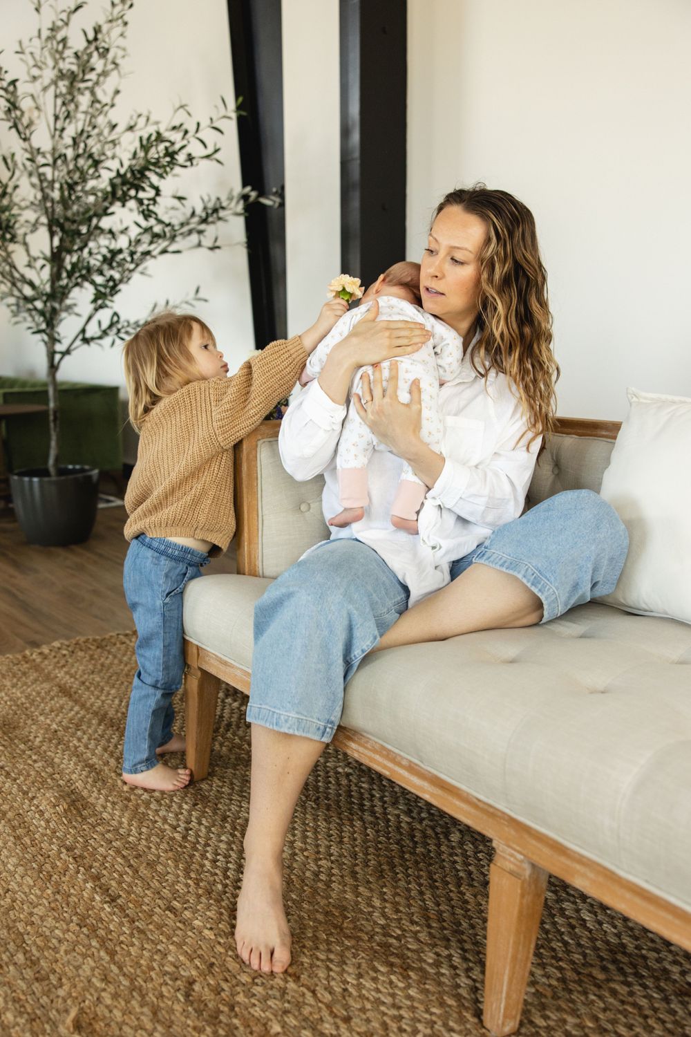 Curly-haired mother holds newborn while toddler in brown sweater reaches out on sofa.