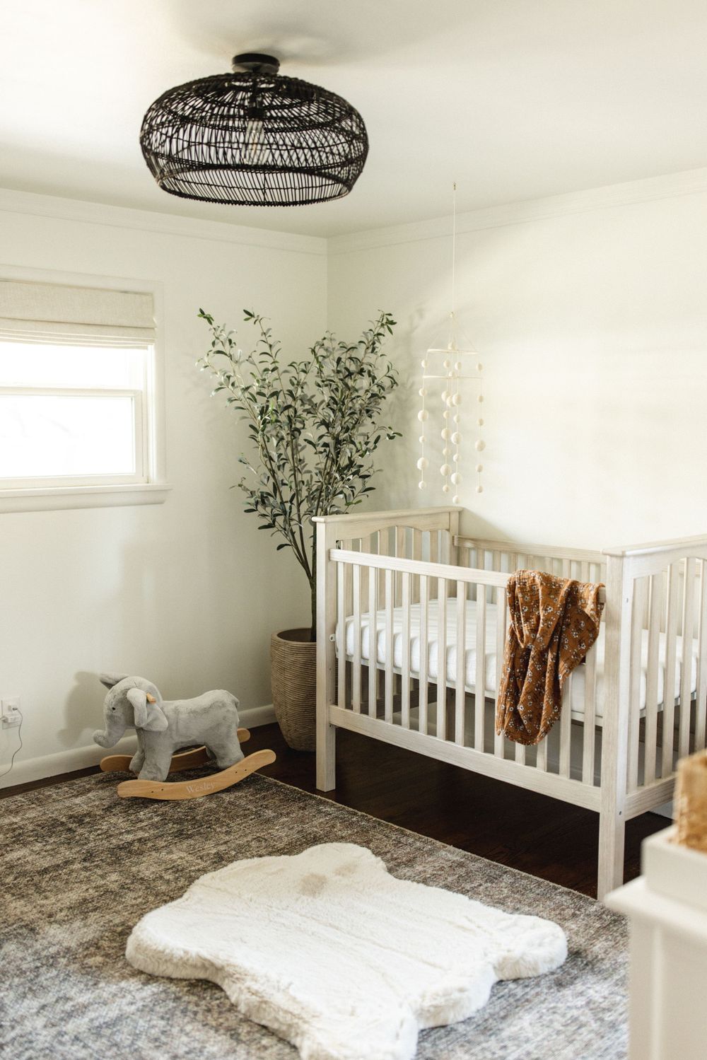 Bright neutral nursery with white crib, rocking horse, faux olive tree, and woven pendant light.