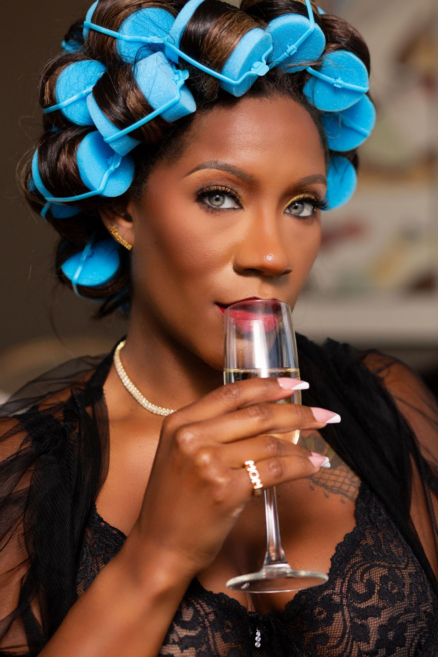 A person wearing bright blue hair rollers and black lace drinks from a champagne glass in a glamorous photoshoot series.