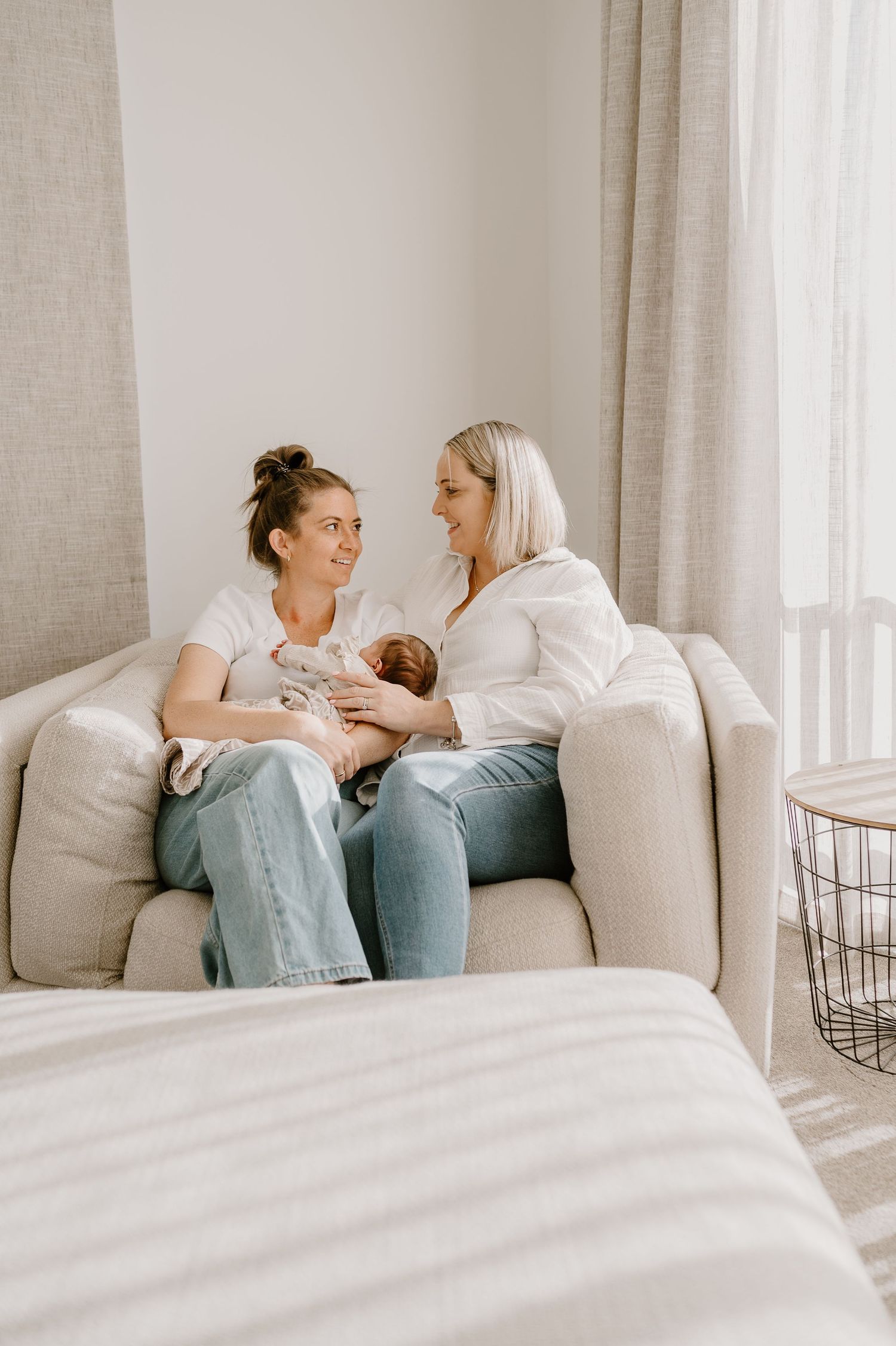 Two people sitting on a couch share an intimate moment with a baby in a bright, minimalist living room setting.