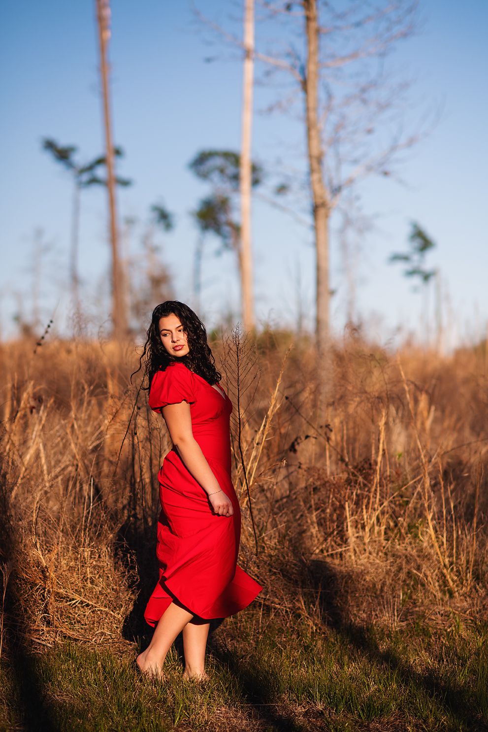 Woman in flowing red dress standing in dry grass field at sunset.