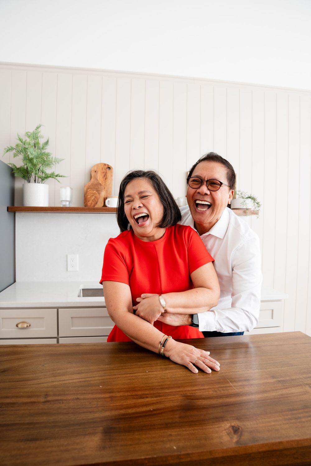A couple laughs together while leaning on a wooden table in a bright kitchen setting.