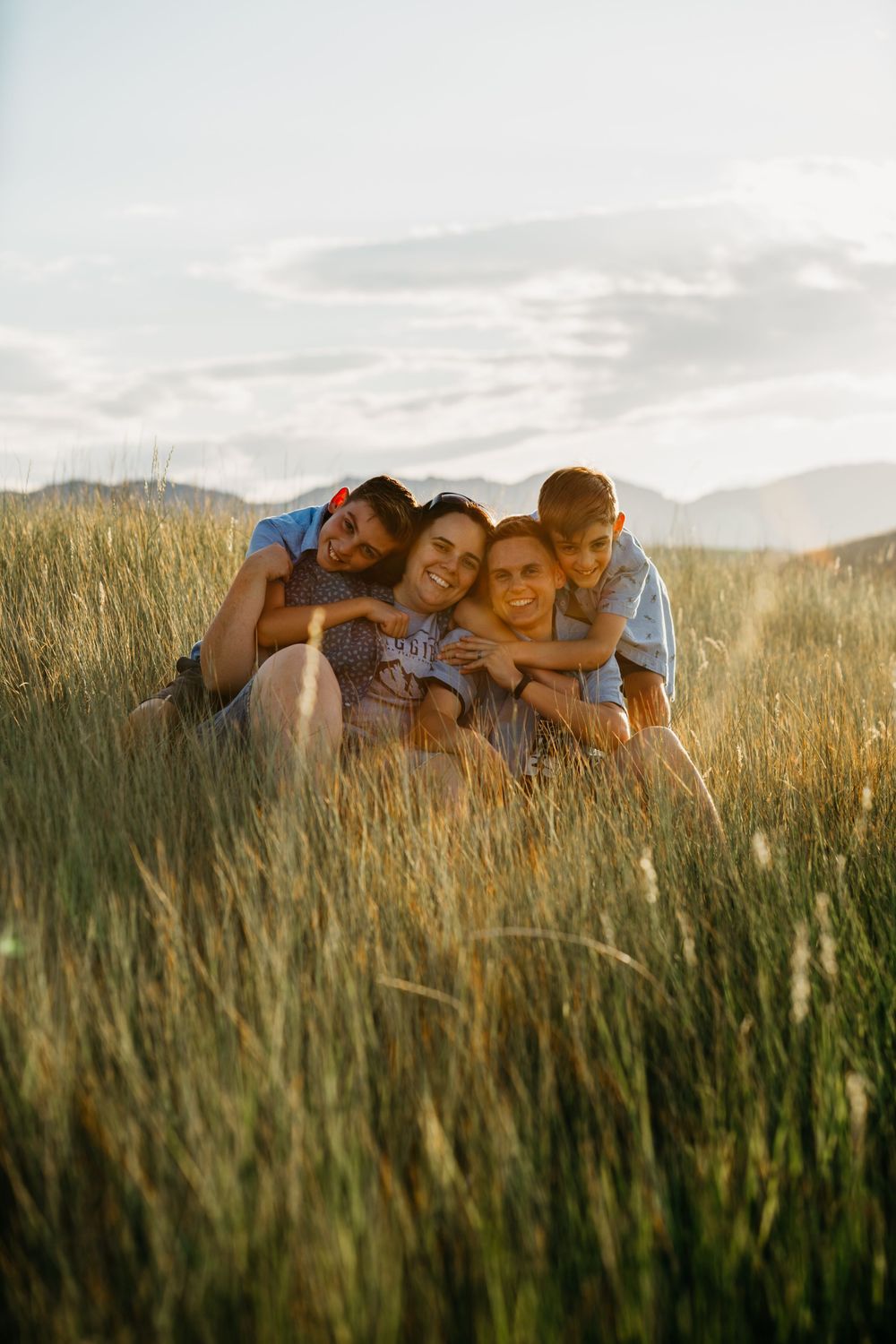 A family cuddles together in tall grass during golden hour with mountains in the background.