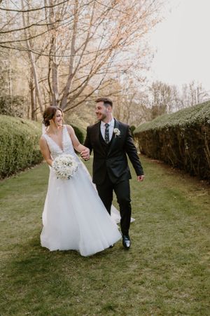 Newlyweds walking and laughing together on a manicured garden path in wedding attire.