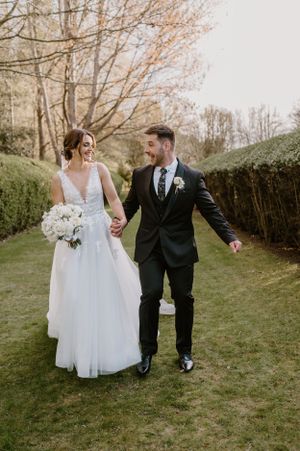 Happy couple walking hand in hand through a garden path in elegant wedding attire.