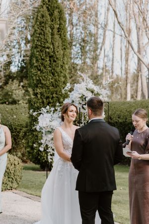 Outdoor wedding ceremony under a floral arch with white blossoms in a garden setting.