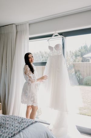 A wedding dress hangs by a window as a person wearing a white lace robe admires it in a bright bedroom setting.