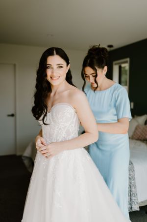 Two people in a bedroom, with one wearing a white lace wedding dress being helped by another in light blue attire.
