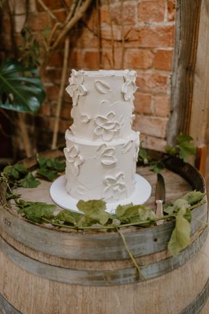 Elegant two-tiered white wedding cake with floral bas-relief details displayed on a rustic wooden barrel.Trents Vineyard, Christchurch photo