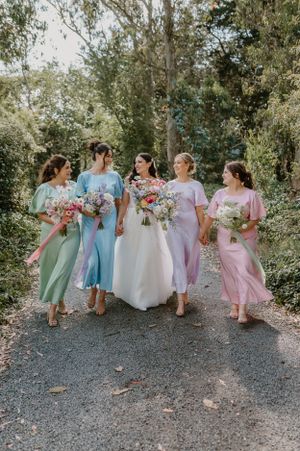 A bridal party in pastel colored bridesmaid dresses walking together on a tree-lined gravel path while holding bouquets. Trents Vineyard