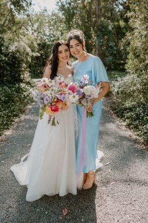 Two people stand together outdoors holding colorful bouquets, one in a white wedding gown and one in blue dress. Trents Vineyard