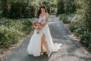 Beautiful bride in flowing white gown with slit holds colorful bouquet while walking on tree-lined garden path. Trents Vineyard