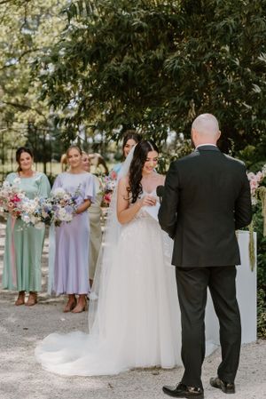 A bride in a white tulle gown exchanges vows with groom as bridesmaids in pastel dresses look on during outdoor ceremony.