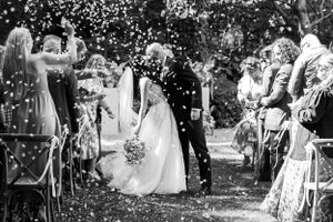 A wedding couple shares a kiss while guests throw confetti during an outdoor garden ceremony celebration.