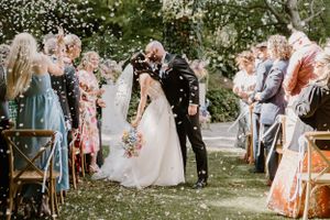 Wedding guests toss confetti over the couple during an outdoor garden ceremony with rustic wooden chairs lining the aisle.