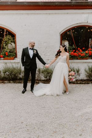A couple celebrates their wedding outdoors by a white brick building adorned with window boxes full of red flowers.