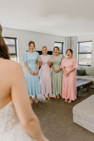Bridesmaids in pastel dresses of blue, pink, sage and lilac stand together in a bright room with ocean views.Trents Vineyard, Christchurch