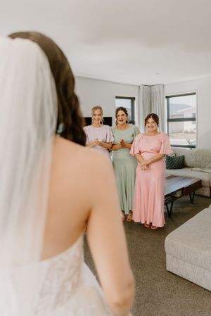 A bride in a white dress faces guests in pastel dresses during an intimate indoor wedding preparation moment.Trents Vineyard, Christchurch