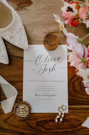 Wedding invitation with white shoes and pink flowers on wooden surface displays names Olivia and Josh.Trents Vineyard, Christchurch photo