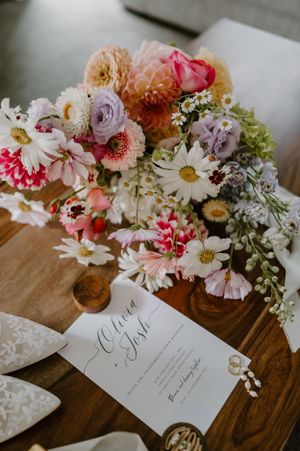 An elegant floral arrangement with daisies and roses sits beside a wedding invitation on a rustic wooden table.